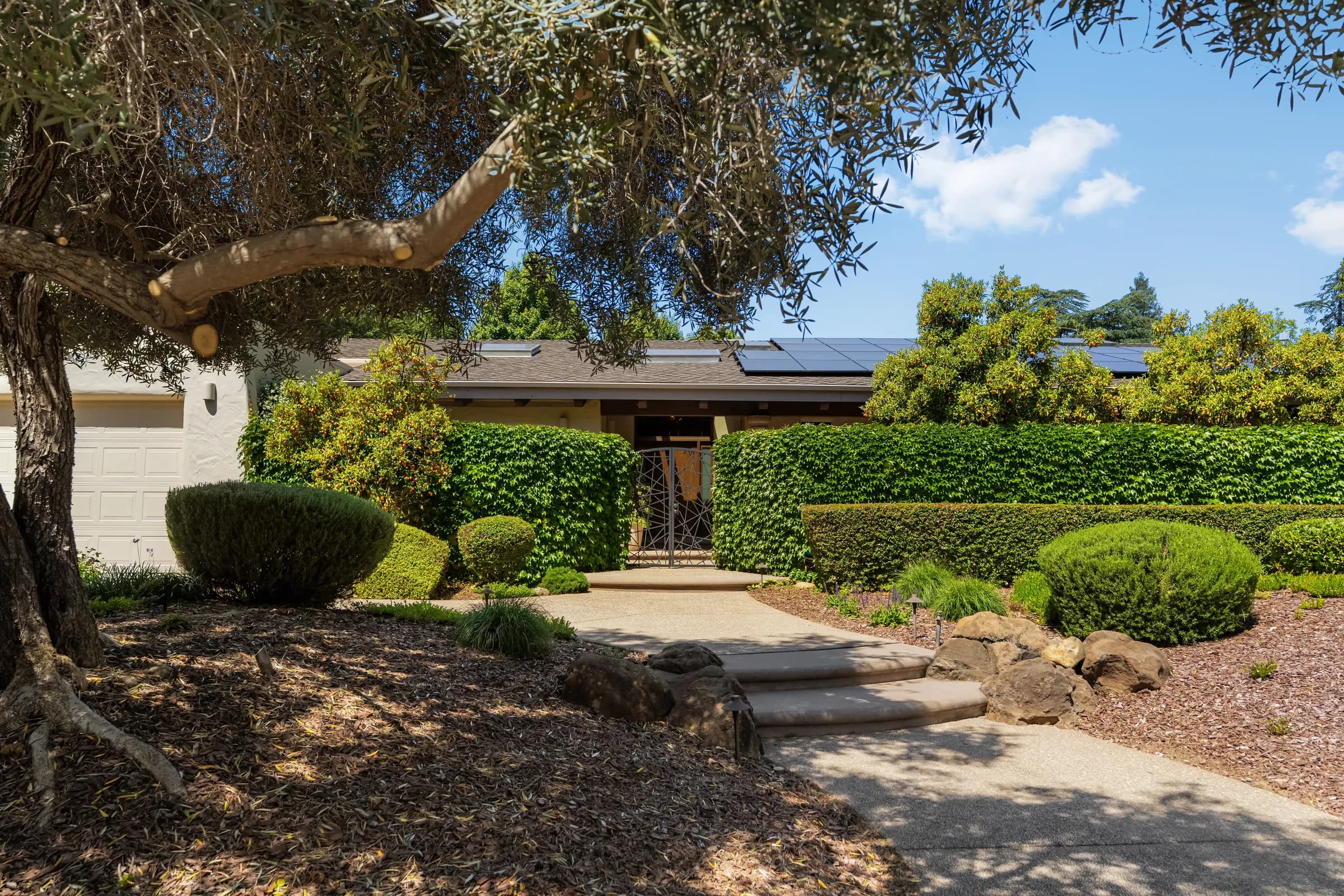 A single-story house behind clipped hedges, with an olive tree, a gravel court and a deep colonnade. Frame 2 of 25.
