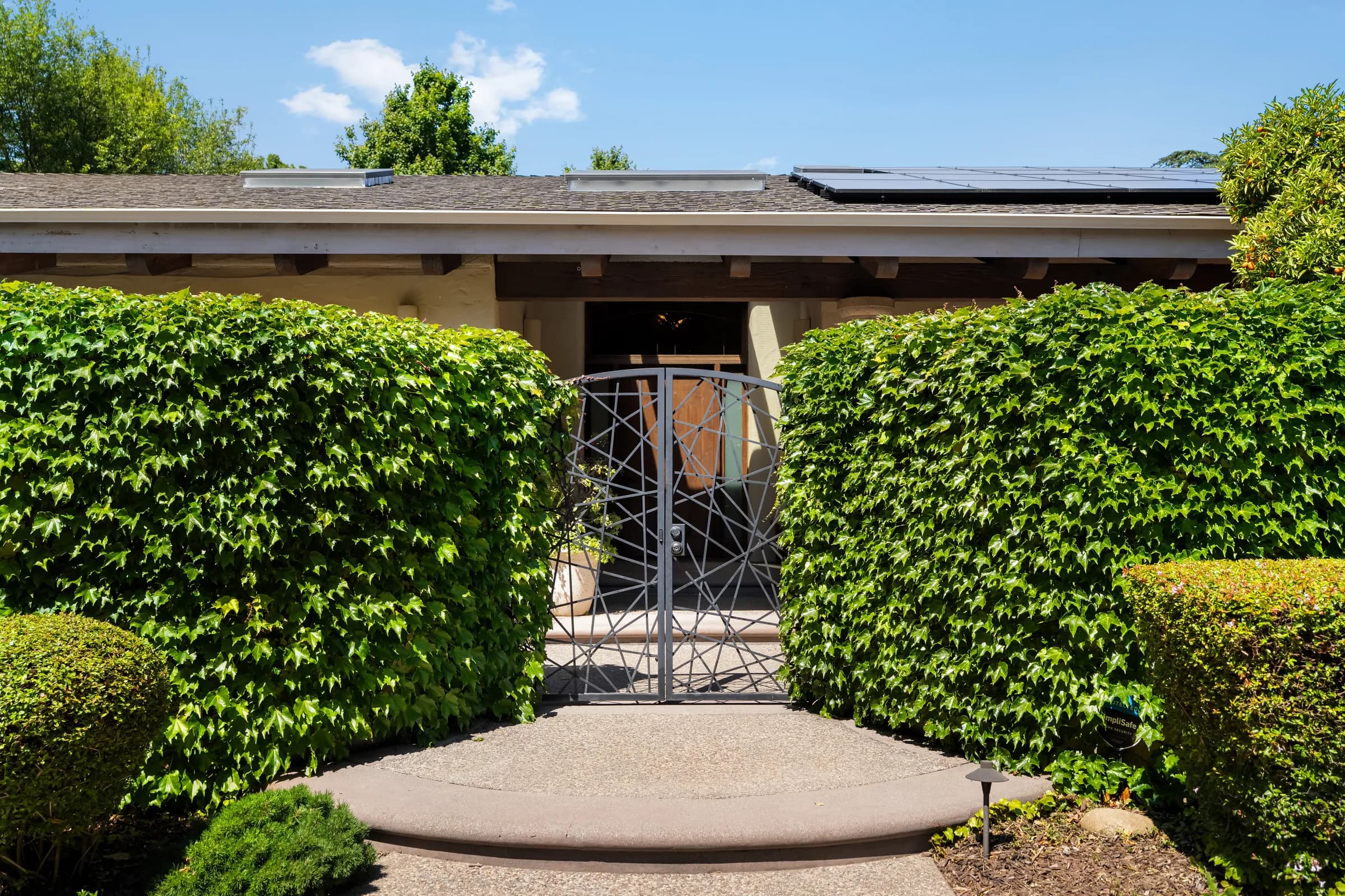 A single-story house behind clipped hedges, with an olive tree, a gravel court and a deep colonnade. Frame 4 of 25.