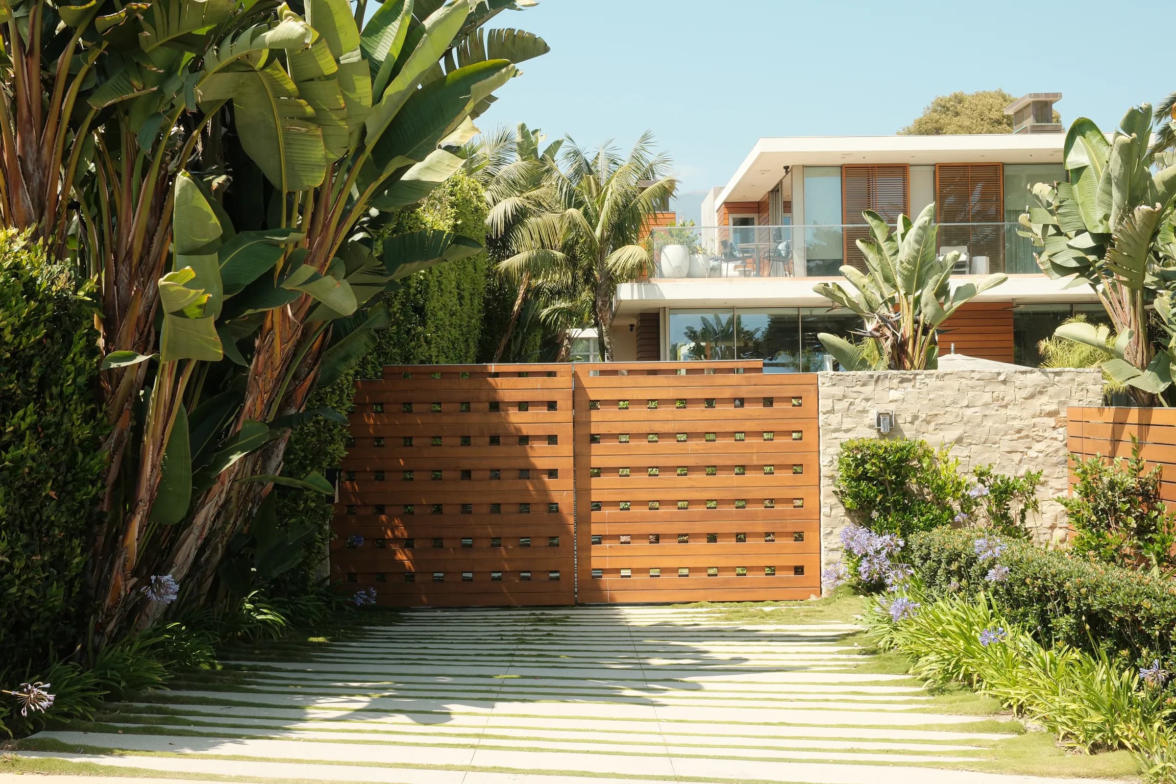A slatted timber gate between mature palms, the house stepping up the slope behind it.