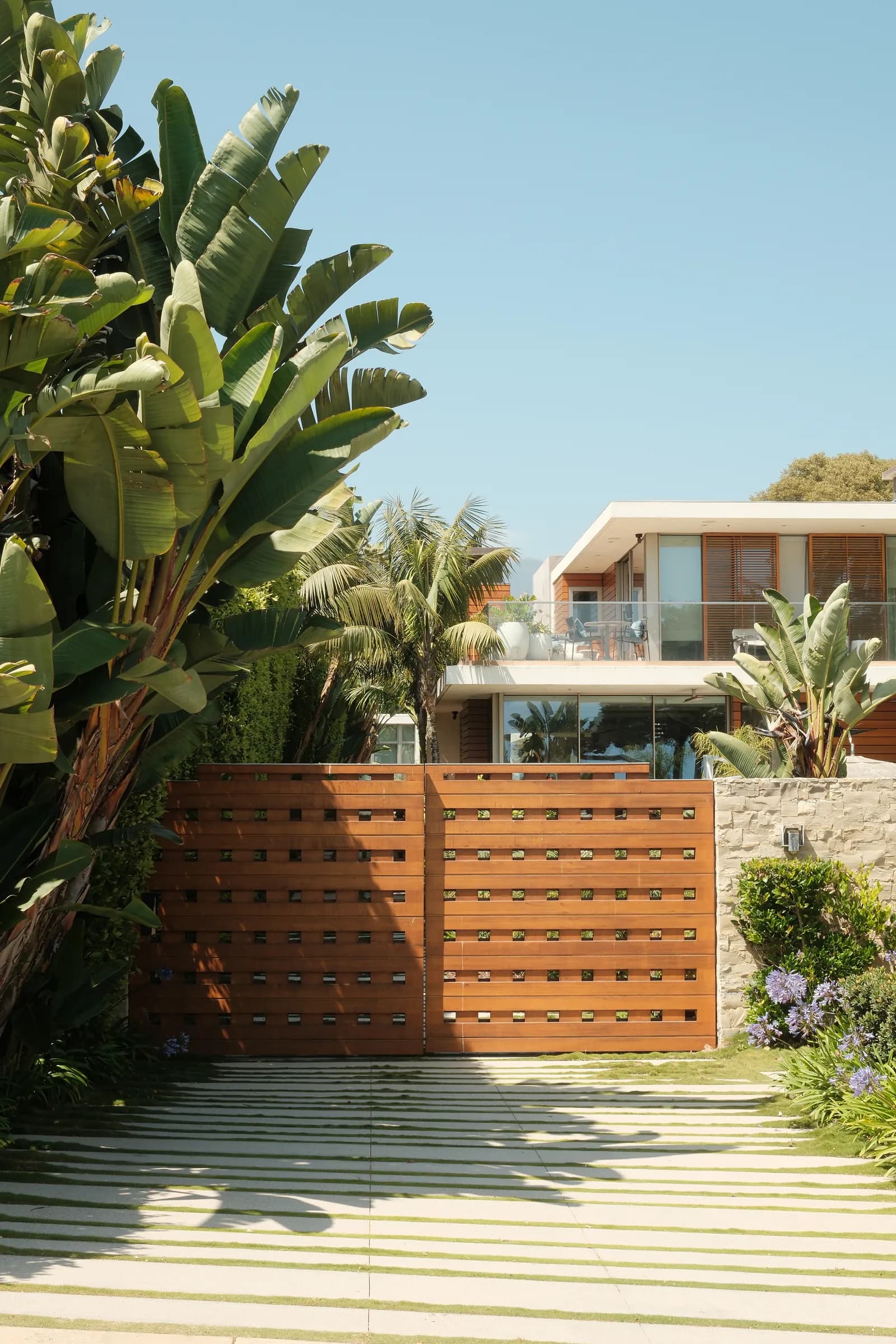 A contemporary house behind a black slatted fence with mature palms, and its interiors: a pale living room, an open kitchen and a detail of a patterned rug against leather. Frame 18 of 23.