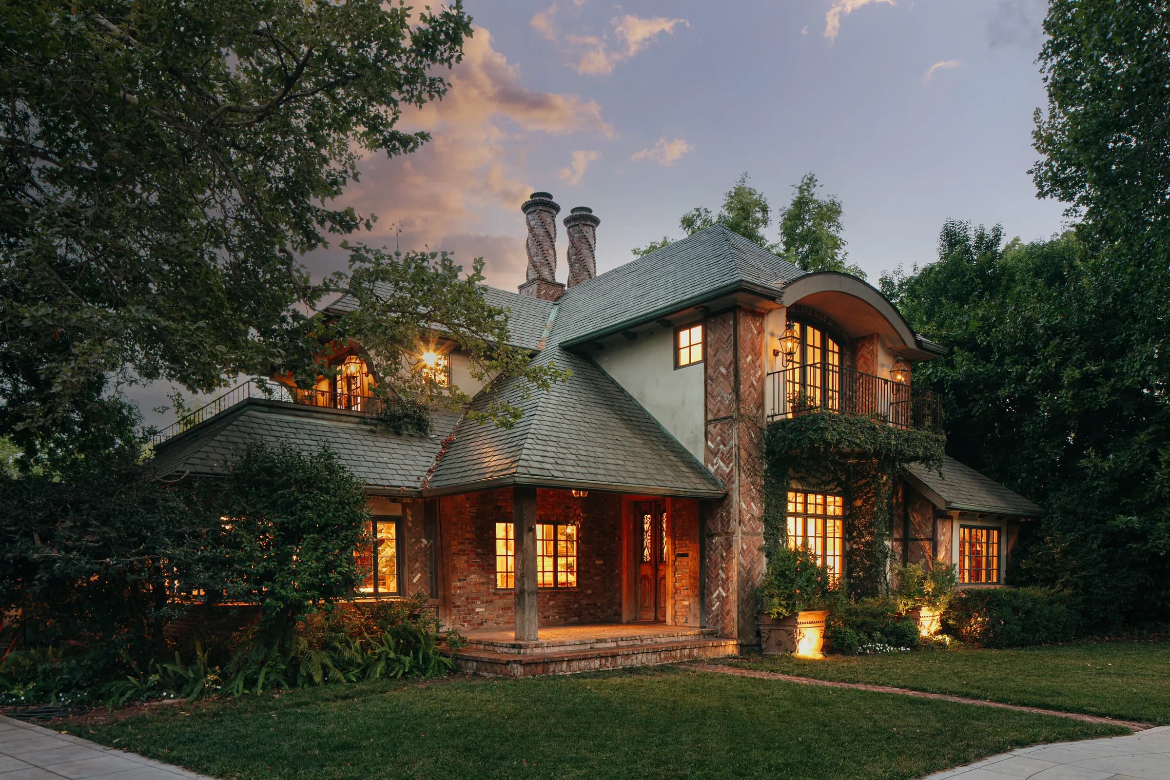 A steep-gabled house at dusk, every window lit warm against a blue sky and a dark lawn.