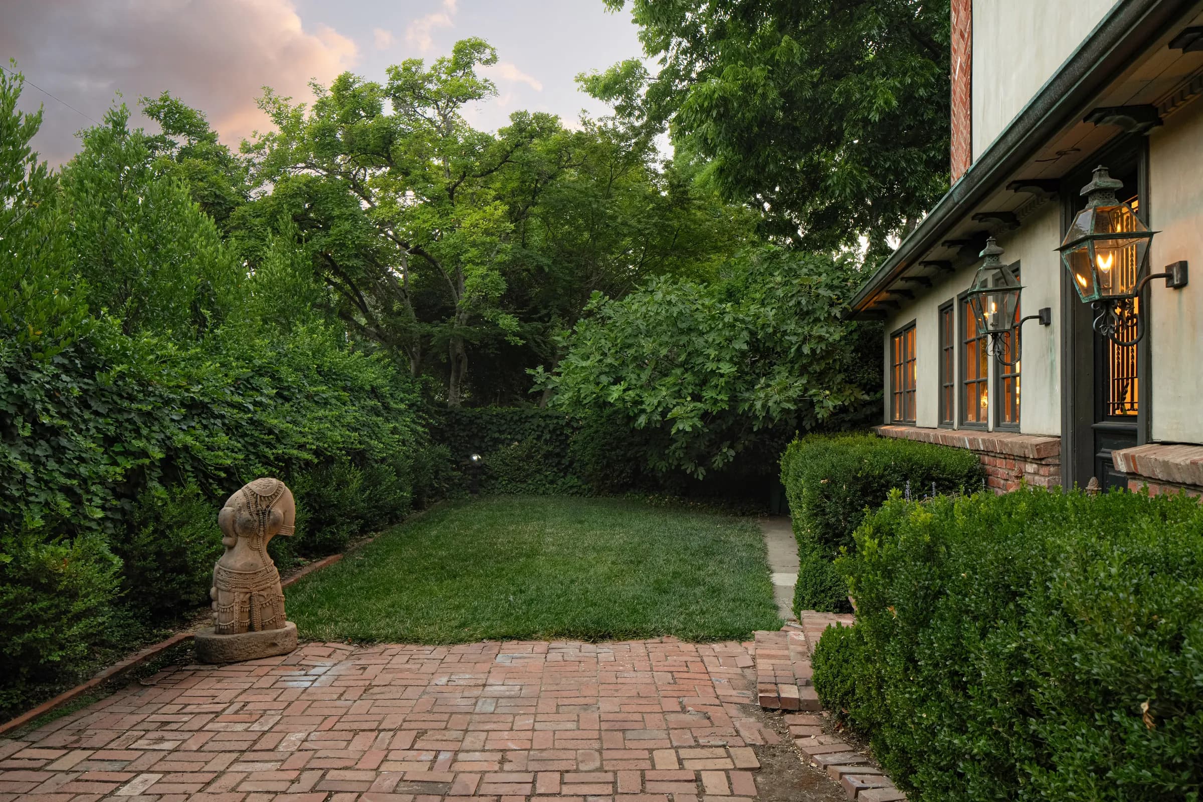 A steep-gabled house photographed at dusk with the interior lights on, its garden, leaded windows and a timber kitchen. Frame 2 of 4.