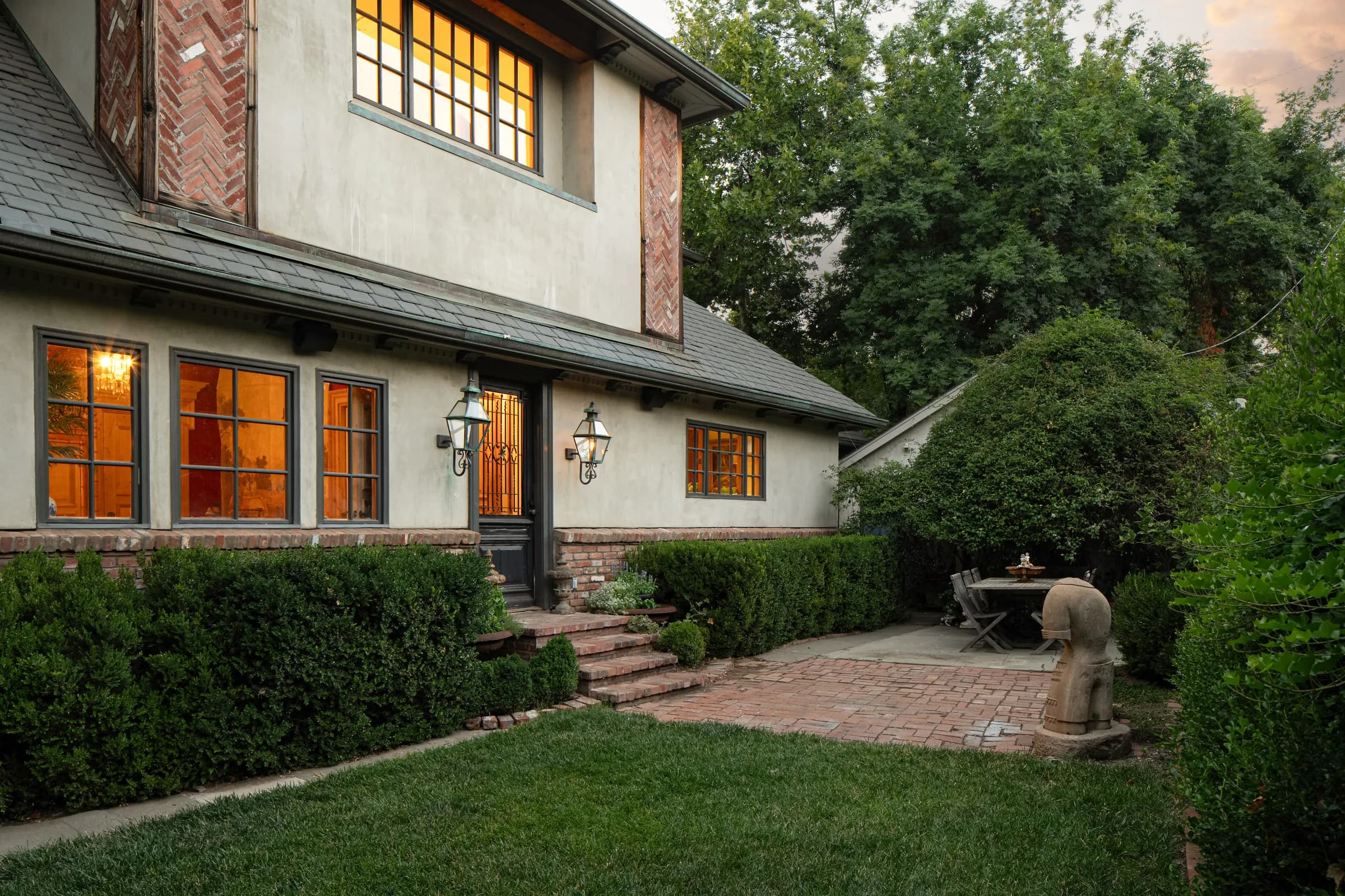 A steep-gabled house photographed at dusk with the interior lights on, its garden, leaded windows and a timber kitchen. Frame 3 of 4.