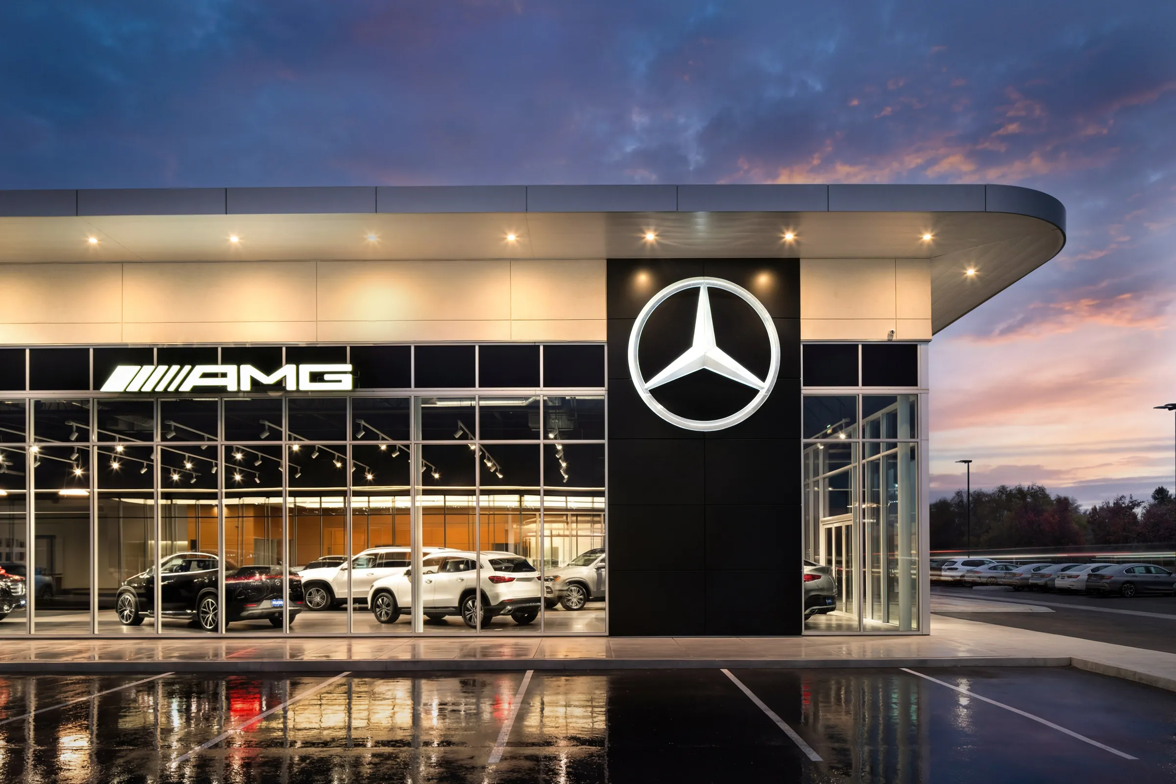 A dealership showroom at dusk, lit from within, its glass frontage reflecting the last of the sky.